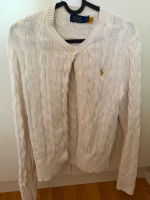 Vit kabelstickad kofta Polo Ralph Lauren - Kabelstickad vit kofta från Polo Ralph Lauren med rund halsringning och knäppning framtill. Klassisk design med ribbade muddar och det ikoniska broderade logot på bröstet. Perfekt till lager på lager och enkel att styla till olika outfits.