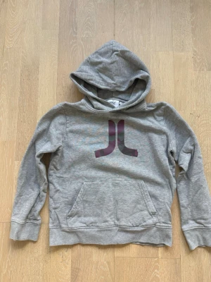 Grå hoodie med JL-logga - Snygg grå hoodie med stor JL-logga i lila på bröstet. Tröjan har huva och en klassisk magficka framtill. Tillverkad i mjukt material och har ribbade muddar vid ärmslut och nederkant. .