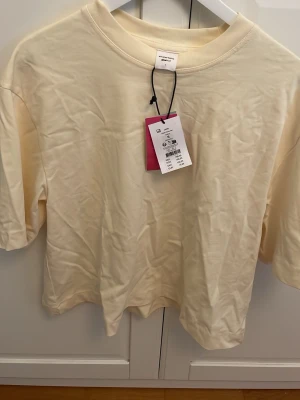 Ljusgul topp fem Gina  - Säljer en enkel och stilren ljusgul topp. T-shirten har korta ärmar, rund halsringning och är gjord i mjuk bomull. Perfekt för dig som gillar en avslappnad och trendig look. Den är helt ny och oanvänd och är skrynklig på bilden men jag stryker den vid köp. Köpte den för 199kr