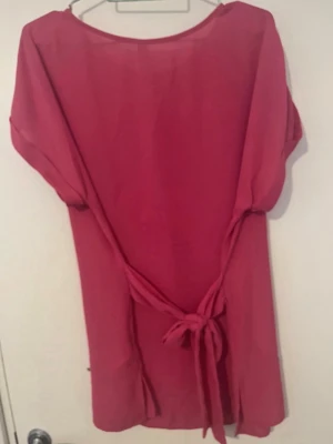 Rosa kortklänning med knytband - Stilren rosa kortklänning med rund hals och korta ärmar. Klänningen har långa knytband i midjan som kan knytas bak för en snygg siluett. Tillverkad i ett lätt material som passar perfekt till varma dagar.