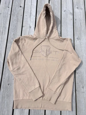 Beige hoodie från GANT med brodyr - Snygg beige hoodie från GANT med broderad logga och sköld på bröstet. Hoodien har dragsko i huvan och ribbade muddar vid ärmslut och nederkant. Tillverkad i mjuk bomullsblandning för extra komfort.