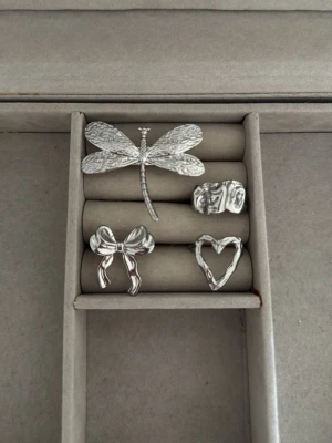 Fyra silverfärgade ringar med motiv - Säljer ett set med fyra unika ringar i silverfärgad metall. Motiven är en trollslända, en rosett, ett hjärta och en ring med abstrakt struktur. Perfekta för dig som gillar detaljerade och lekfulla accessoarer. Justerbara ringar, trollsländan är köpt från Lyla och den abstrakta ringen är köpt här på plick. Ringarna rostar inte🫶🏻