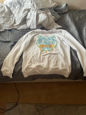 Vit stickad Kenzo tröja med tiger - Säljer en vit stickad tröja från Kenzo med det ikoniska tigerhuvudet broderat i blått, grönt och orange framtill. Tröjan har rund halsringning och ribbade muddar. Perfekt för dig som vill ha en statement piece i garderoben.               Priser kan diskuteras 