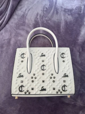 Vit handväska med nitar och detaljer - Säljer en vit handväska från Christian Louboutin med silverfärgade nitar och logodetaljer på framsidan. Väskan har två handtag, fyrkantig form och röda sidopaneler med präglad logga. Tillverkad i läder med metallfötter undertill.