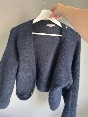Mörkblå stickad kofta från Gina - Mysig mörkblå kofta med öppen front och lång ärm. Koftan är fluffig och har en mjuk, borstad yta som ger en avslappnad look. Perfekt att slänga över en t-shirt eller topp för extra värme. Enkel och stilren design med knappar. Använd 1 gång