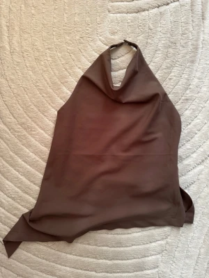 Brun halterneck topp i satin - Säljer en brun halterneck topp i mjukt satinliknande material. Toppen har en asymmetrisk form och knäpps i nacken. Perfekt för dig som gillar minimalistisk och trendig stil.