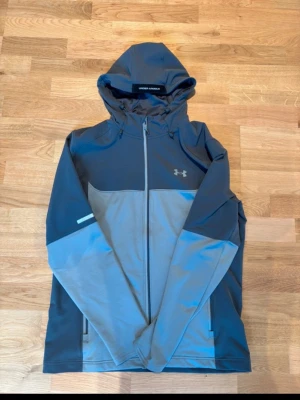 Grå Under Armour vindjacka - Snygg tvåfärgad vindjacka från Under Armour i mörkgrått och ljusgrått. Jackan har huva, hel dragkedja framtill, två sidofickor med dragkedja och reflekterande detalj på ärmen. Perfekt för blåsiga dagar och sportiga outfits. OBS PRISET KAN DISKUTERAS!