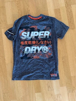 Superdry grå t-shirt med tryck - Cool grå t-shirt från Superdry med stort tryck framtill i vitt, orange och svart. T-shirten har korta ärmar och rund hals. Perfekt för dig som gillar streetwear och vill sticka ut med en snygg logga och japanska tecken.