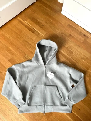 Saint Alexander zip-up Hoodie  - Snygg ljusgrå hoodie från Saint Alexander med dragkedja framtill och stor känguruficka. Hoodien har en rejäl huva och ribbade muddar vid ärmslut och nederkant. Perfekt för en chill och avslappnad stil. Materialet känns mjukt och bekvämt.