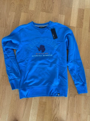 Blå sweatshirt från Sail Racing - Blå sweatshirt från Sail Racing med tryck på bröstet där det står 'SAIL RACING' och 'ANTARCTICA EXPEDITION'. Tröjan har rund halsringning, ribbade muddar och är tillverkad i mjukt bomullsmaterial. Perfekt för en avslappnad och sportig stil.