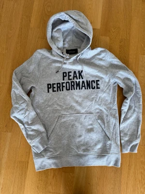 Grå hoodie Peak Performance M - Snygg grå hoodie från Peak Performance med stor svart logga på bröstet. Klassisk känguruficka framtill, dragsko i huvan och mjukt, borstat foder på insidan. Perfekt för chill dagar eller när du vill ha en sportig look.