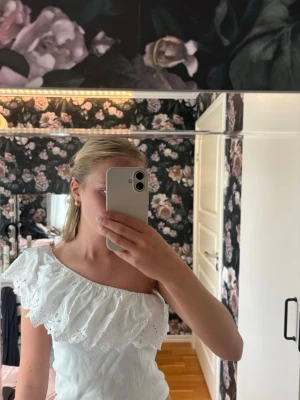 Vit oneshoulder topp med volang - Superfin vit topp med oneshoulder och bred volangkant i broderad spets. Toppen är ärmlös på ena sidan och har en smal axelrem på andra. Perfekt till sommaren 🌸