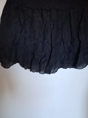 Svart volangkjol med resårmidja - Kort svart kjol med volang och resår i midjan. Kjolen har ett tunt, lätt material och en subtil struktur i tyget som ger extra detalj. Perfekt för dig som gillar en enkel men ändå trendig look.