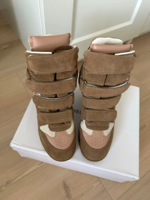 Bruna höga sneakers med kardborreband - Snygga höga sneakers i mocka och läder med flera kardborreband. Skorna är bruna med beige och ljusrosa detaljer samt har en chunky sula. Perfekta för dig som gillar streetstyle och vill sticka ut med en unik design.