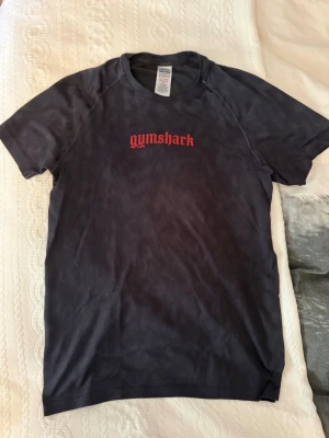 Svart Gymshark t-shirt  - Snygg svart t-shirt från Gymshark med röd logga på bröstet. T-shirten har korta ärmar och en klassisk passform. Perfekt för dig som gillar stilrena träningskläder och vill ha något bekvämt till gymmet.