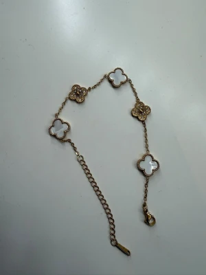 Guldfärgat armband med fyrklöver - Snyggt armband i guldfärg med fem fyrklöverformade länkar, varav tre har vit pärlemor och två är dekorerade med små gnistrande stenar. Kedjan är tunn och har ett justerbart lås. Perfekt accessoar för dig som gillar detaljer och elegant design.