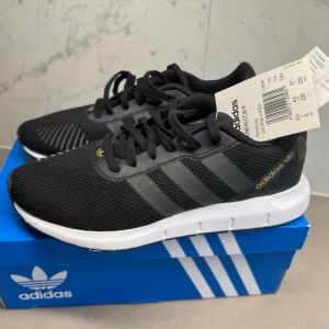 Svarta Adidas sneakers med mesh - Snygga svarta sneakers från Adidas med klassiska tre ränder i mörkgrått på sidorna. Ovandelen är i ventilerande mesh och skon har vita sulor för en fräsch kontrast. Diskreta gulddetaljer på plösen och sidan. Perfekta för en sportig och trendig look.