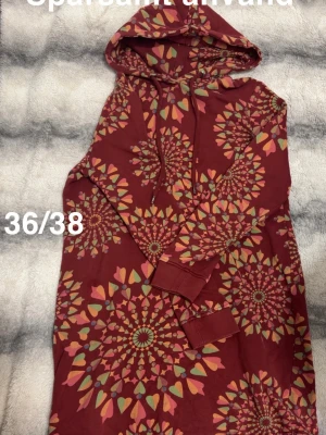 Vinröd klänning med mönster - Vinröd klänning med huva och långa ärmar, dekorerad med färgglada mandala-liknande mönster i rosa, gul och grön. Klänningen har en avslappnad passform, perfekt för dig som gillar unika prints.🌸 Sparsamt använd. Storlek 36/38