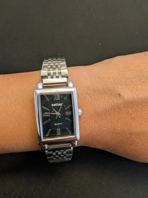Rektangulär silverfärgad klocka - Stilren klocka med rektangulärt svart urtavla och silverfärgat metallarmband. Urtavlan har vita markeringar och romerska siffror vid 12 och 6. Klassisk design som passar till många stilar.