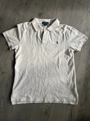 Vit pikétröja från Polo Ralph Lauren - Klassisk vit pikétröja från Polo Ralph Lauren med blå logga broderad på bröstet. Tröjan har korta ärmar, krage och knappar framtill. Tillverkad i bomull för en skön och luftig känsla. Perfekt till jeans eller shorts. Pikén är i mycket bra skick och kan strykas så den inte ser så skrynklig ut:)