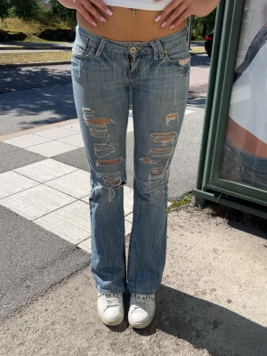 Ljusblå slitna bootcut jeans - Säljer ett par ljusblå bootcut jeans med flera stora slitningar och hål framtill på båda benen. Jeansen har låg midja, klassiska fem fickor och är tillverkade i denim. Perfekta för en avslappnad och trendig look. Står något konstigt märke i byxorna om ni vill att jag ska ta kort på de.