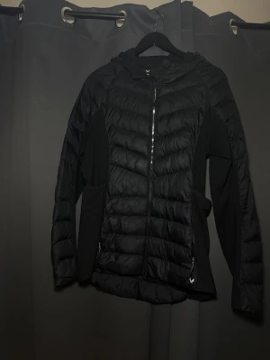 Svart pufferjacka med huva - Snygg svart pufferjacka med huva och dragkedja framtill. Jackan har quiltade partier och släta sidopaneler för en sportig look. Två fickor med dragkedja och längre passform baktill. Perfekt för kalla vinterdagar. Märket är SOC. Relativt använd, lite slitage, se bilder.