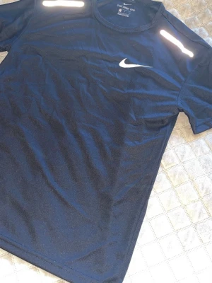 Svart Nike t-shirt med reflexdetaljer - Snygg svart t-shirt från Nike med vita reflexdetaljer på axlarna och klassisk Nike-logga på bröstet. Tillverkad i ett lätt och funktionellt material som passar perfekt till träning. Rund halsringning och korta ärmar för en sportig look.
