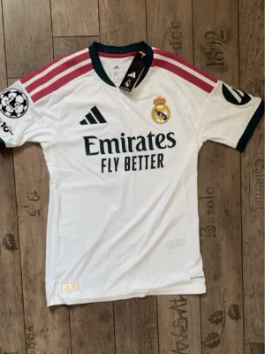 Real Madrid matchtröja Vini Jr. 7 - Säljer en vit Real Madrid matchtröja från Adidas med Vini Jr. och nummer 7 på ryggen. Tröjan har rosa och svarta detaljer, klubbmärke och sponsortryck framtill samt Champions League-märke på ärmen. Perfekt för fotbollsfans!