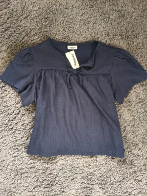Marinblå blus med knyt i bomull - Söt marinblå blus från Kappahl med korta puffärmar och en bred krage med knyt framtill. Tillverkad i mjuk bomull som känns skön mot huden. Perfekt för dig som gillar en enkel men ändå trendig look.