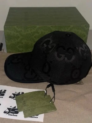 Svart keps med GG-mönster från Gucci - Snygg svart keps från Gucci med det klassiska GG-mönstret i svart ton-i-ton. Kepsen har böjd skärm och justerbar rem bak. Tillverkad i ett slitstarkt tygmaterial som ger en exklusiv känsla. Perfekt för dig som vill ha en stilren accessoar med lyxig touch.