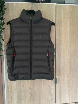 Svart dunväst från Polo Ralph Lauren - Snygg svart dunväst från Polo Ralph Lauren med hög krage och två dragkedjeförsedda fickor. Västen har en diskret logga på bröstet och quiltad design som ger en modern look. Perfekt för lager-på-lager och håller dig varm under kyliga dagar.västen är ganska liten i storleken så du vet.