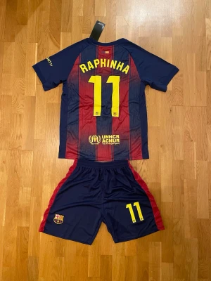 Raphinha FC Barcelona hemmaställ 11 - Säljer ett FC Barcelona matchställ med tröja och shorts. Tröjan har blå och röda ränder, klubbmärke, Spotify-logga och Raphinha 11 på ryggen. Shortsen är blå med röda detaljer och nummer 11. Perfekt för fotbollsträning eller att heja på laget.