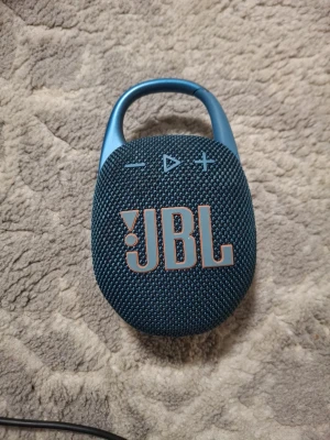 Blå JBL Clip bärbar högtalare - Snygg blå JBL Clip bärbar högtalare med robust metallklämma och tygfront. Smidig oval form och tydliga knappar för volym och uppspelning på ovansidan. Perfekt för att ta med sig musiken överallt tack vare den smarta karbinhaken.