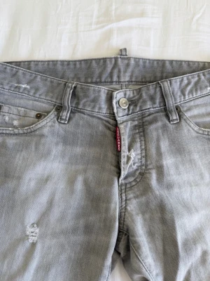DSQUARED2 jeans - Säljer ett par grå jeans med raka ben och slitna detaljer. Jeansen har klassisk femficksmodell, knappgylf och en röd etikett vid fickan. Materialet är denim i bomull och de har en avslappnad vibe. Äktighets bevisade av Sellpy 