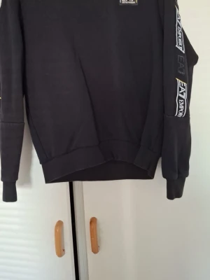 Svart hoodie från EA7 Emporio Armani - Snygg svart hoodie från EA7 Emporio Armani med logga på bröstet och längs ena ärmen. Klassisk passform med ribbade muddar och mjukt material. Perfekt för en avslappnad och trendig look.