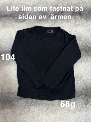 Svart långärmad topp  - En enkel svart långärmad topp med rund halsringning och diskret spetskant., perfekt för en clean och stilren look. Passar till det mesta och är lätt att matcha med andra plagg.🌸 Lite lim som fastnat på sidan av ärmen. Storlek 104 