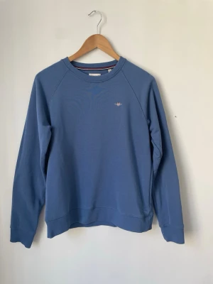 Blå sweatshirt från Gant - Säljer en stilren blå sweatshirt från Gant med rund halsringning och diskret broderad logga på bröstet. Tröjan har långa ärmar och är gjord i mjukt bomullsmaterial. Perfekt för en avslappnad och snygg look. Tröjan är i väldigt bra skick och bara använd ett fåtal gånger.