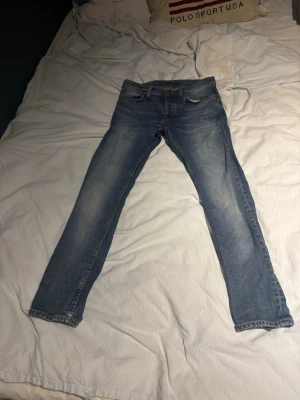 Ljusblå jeans från Nudie Jeans - Säljer ett par ljusblå jeans från Nudie Jeans med klassisk femficksmodell och raka ben. Jeansen har snygga slitningar och kontrastsömmar. Perfekta för dig som gillar en avslappnad och stilren look.
