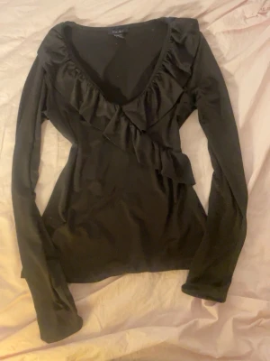 Svart volangtopp från New Yorker - Säljer en svart långärmad topp från newyorker med snygg volangdetalj längs den djupa v-ringningen. Toppen har en feminin och figurnära passform som passar perfekt till jeans eller kjol.
