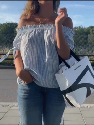 Blårutig offshoulder topp - Söt offshoulder topp i ljusblått och vitt med rutigt mönster. Toppen har resår vid axlarna och knytband på ärmarna för extra detalj. Perfekt till jeans eller kjol för en fräsch sommarlook.