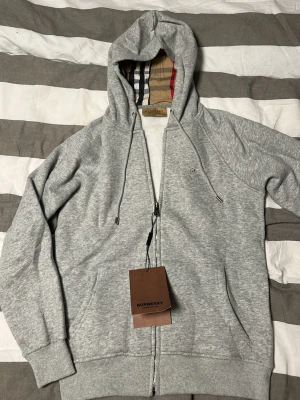 Grå zip-hoodie från Burberry - Snygg grå hoodie från Burberry med dragkedja, huva och klassiskt rutigt foder i luvan. Mjuk bomullsblandning, fickor framtill och diskret Burberry-logga på bröstet. Perfekt för en chill och stilren look. 