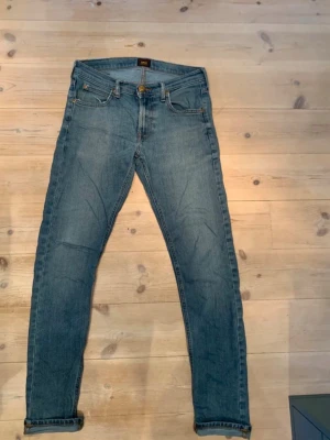 Blå slim jeans från Lee - Snygga blå slim jeans från Lee med klassisk femficksdesign och uppvikta benslut. Jeansen har en mellanblå tvätt och Lee-logga på bakfickan. Perfekta för en avslappnad och trendig stil.