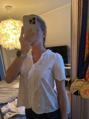Vit blus med broderade detaljer - Söt vit blus med korta ärmar och stora broderade volangdetaljer längs ringningen. Blusen har ett luftigt tyg med hålbroderier och en avslappnad passform. Perfekt för en trendig och romantisk look. Köpt från Missay🌸