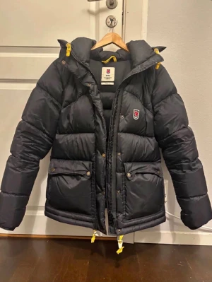 Fjällräven expedition lite Women S - Den är använd en vinter och är precis som ny. Den har inga deffekter. Snygg svart dunjacka från Fjällräven med gulddetaljer och gula snören. Jackan har två stora fickor framtill, dragkedja och tryckknappar, samt en huva. Fjällräven-logga på bröstet och vadderad design för extra värme. Storlek S 