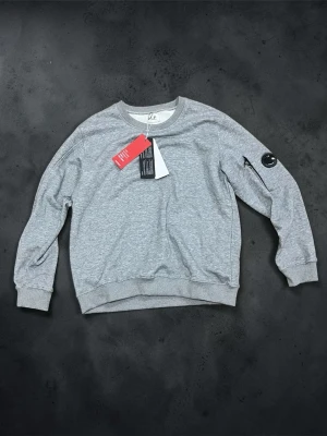 Grå sweatshirt med patch på ärmen - Stilren grå sweatshirt med rund halsringning och ribbade muddar vid ärmslut och nederkant. På vänster ärm finns en svart patch med smiley. Perfekt för en avslappnad och trendig look.
