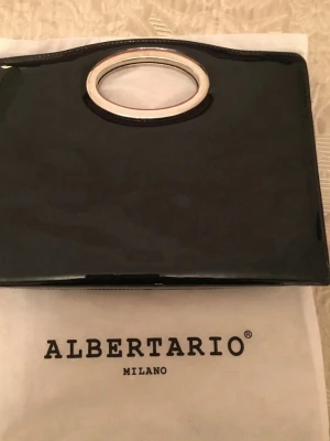 Svart lackad kuvertväska Albertario - Stilren svart kuvertväska i lackat material från Albertario Milano. Väskan har en oval metallring som handtag och en elegant, glansig finish. Perfekt accessoar för att lyfta din outfit med en modern touch.