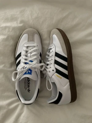 Adidas Samba vita och svarta sneakers - Helt oanvända klassiska Adidas Samba sneakers i vitt och svart med bruna sulor. Skorna har tre svarta ränder på sidan, vit ovandel i läder och detaljer i grå mocka vid tån. Guldtexten 'SAMBA' syns på sidan. Perfekta för en sportig och stilren look.