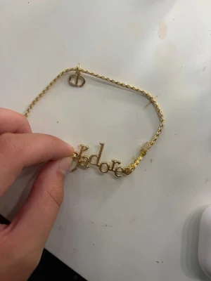 Guldigt J'adore armband med flätad band - Snyggt armband med flätad vit och guldig tråd samt metalltexten 'J'adore' i guld. Armbandet har även en liten hängande detalj med bokstäverna CD i metall. Perfekt accessoar för att lyfta din stil.