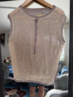Beige stickad ärmlös topp med knappar - Säljer en beige och brun stickad ärmlös topp med rund hals och dekorativa knappar framtill. Toppen har ett rutigt mönster och ribbade kanter i metallicbrunt. Perfekt för dig som gillar retrovibbar och unika detaljer.