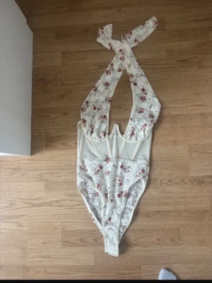 Vit spetsbody med blommönster - Säljer en vit body i spets med blommönster i rött och grönt. Bodyn har djup urringning, breda axelband och transparent meshdetalj vid midjan. Materialet är stretchigt och mjukt, perfekt för dig som gillar romantisk och feminin stil.